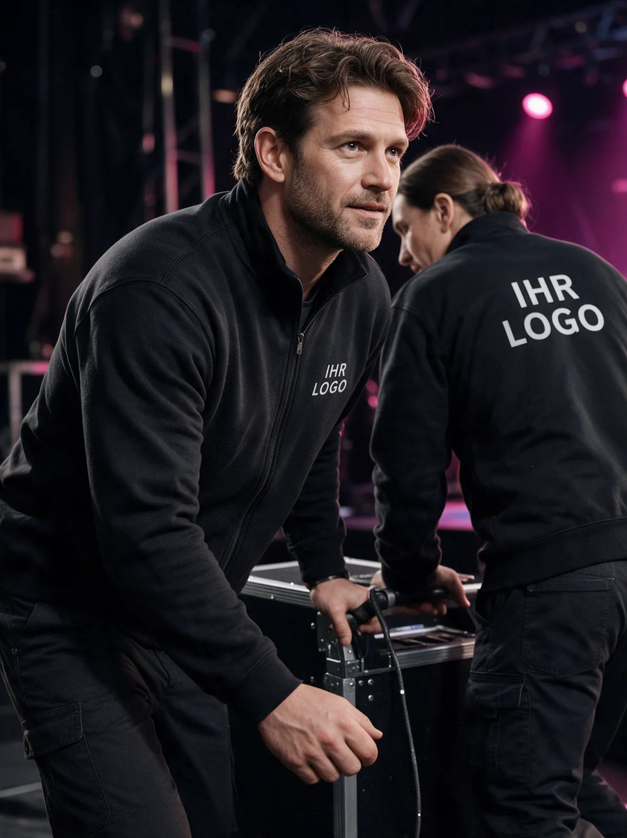 Eventcrew in schwarzen Sweatjacken mit Logoaufdruck bewegt einen Transportkoffer hinter der Bühne.